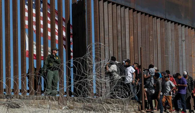 Migrantes miran a través de la valla mientras un agente de la Patrulla Fronteriza vigila - Cruce fronterizo de El Chaparral en Tijuana, México.